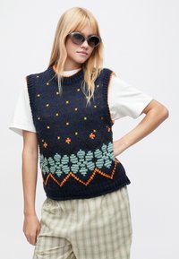 Sweter dzianinowy bez rękawów w kolorze granatowym z pomarańczowo-zielonymi wzorami kwiatowymi, z prążkowanym kołnierzem i ściągaczem, noszony na białej koszulce.