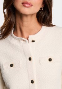 Pullover crema con bottoni e finitura testurizzata, caratterizzato da dettagli neri sui bottoni e due tasche sul petto. Collo rotondo semplice.