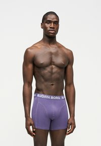 Μυώδης άνδρας με σκούρο δέρμα, όρθιος, φορώντας μοβ μποξεράκια Björn Borg, μπροστά σε απλό φωτεινό φόντο.