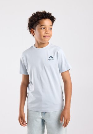 Jongen met krullend haar die een lichtblauw "Into the Waves" t-shirt en lichtgewassen jeans draagt en met een lichte glimlach naar rechts kijkt