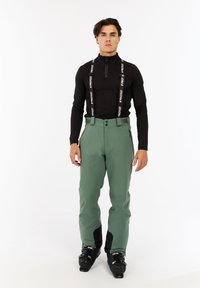 Skibroek in gedempt groene stof met zwarte accenten, voorzien van verstelbare bretels, een ritssluiting aan de voorkant en een taps toelopende pasvorm. Zwarte skibotten maken de look compleet.