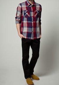 Man som bär en röd, vit och blå rutmönstrad skjorta med knappar, svarta byxor och senapsgula mockaskor, står med händerna i fickorna.