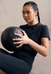 Fekete fitneszlabda, amelyet egy fitted fekete Nike pólót és sötét leggingset viselő személy tart a kezében, copfba font hajjal, semleges háttér előtt.