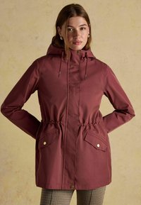 Burgundowa kurtka przeciwdeszczowa z kapturem, z wąskim pasem, kieszeniami i zatrzaskami. Gładka struktura z lekkiego, wodoodpornego materiału.