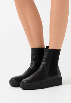 Botas de tobillo de cuero negro con paneles laterales elásticos, suela texturada negra y una lengüeta en la parte posterior para ponérselas fácilmente. Acabado suave.