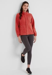 Fleece jacket in coral with a high collar, front buttons, and side pockets; paired with dark leggings and black athletic shoes.