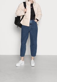 Persona vistiendo una chaqueta acolchada beige claro, camiseta negra, jeans de cintura alta azules, zapatillas blancas y llevando una mochila negra sobre un hombro.