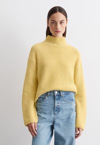 Gelber, gerippter Rollkragenpullover mit weiten Ärmeln, kombiniert mit hoch geschnittenen hellblauen Jeans, die eine lockere Passform und Textur zeigen.