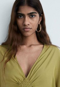 Olivgrön blus med V-ringning och rynkdetaljer i mitten. Modellen bär stora guldfärgade hoops. Slätt, lätt material.