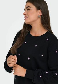 Schwarzer Sweatshirt mit verstreuten, pinken Herz-Stickereien. Mit Rundhalsausschnitt und gerippten Bündchen, getragen von einem Model, das Schmuck präsentiert.