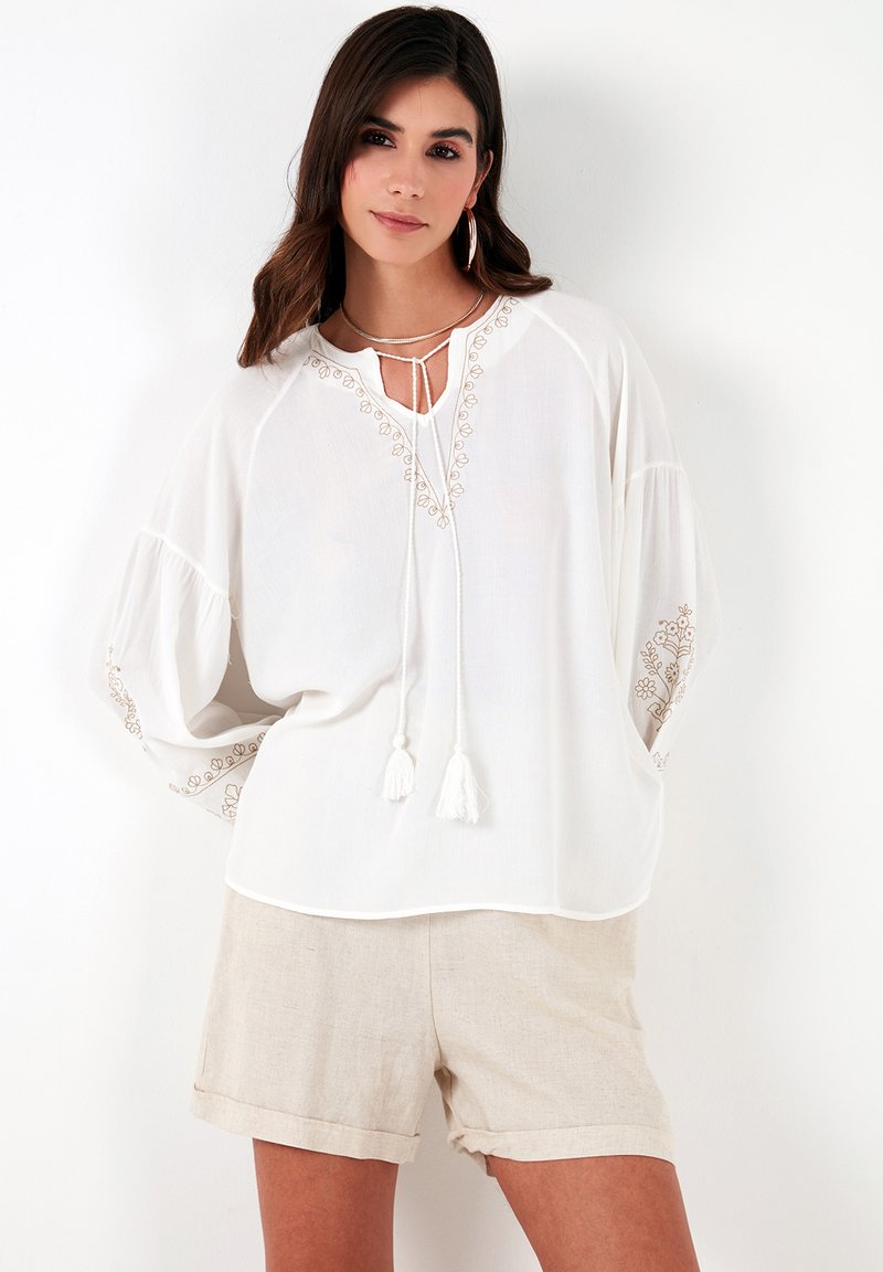 White blouse with floral embroidery along the neckline and sleeves, featuring a loose fit and tied drawstring with tassels. Linen shorts.