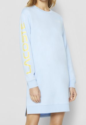 Vrouw die een lichtblauwe sweatshirtjurk met lange mouwen en gele "Lacoste" tekst op de rechtermouw draagt, staand tegen een effen achtergrond.