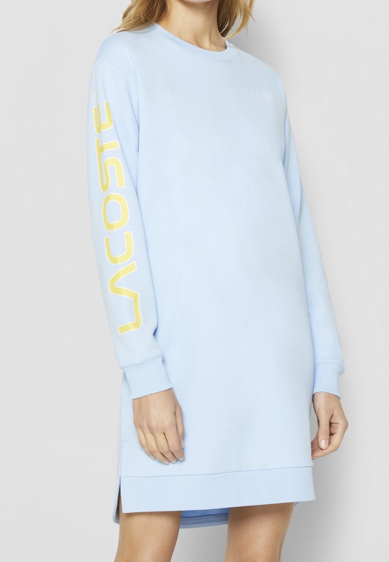 Femme portant une robe sweat-shirt bleu clair à manches longues avec le texte jaune « Lacoste » sur la manche droite, debout devant un fond uni.