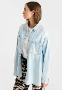 Lichtblauwe denim shirt met een knoopsluiting aan de voorkant, oversized pasvorm, twee borstzakken en subtiel logo-detail op de linkse zak.