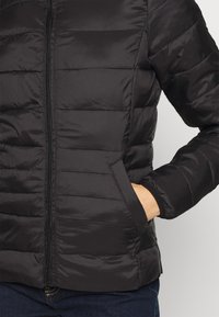 Svart uppblåsbart jacka med dragkedja framtill, horisontell sömnad och sidofickor. Tillverkad av smidigt, vattenavvisande tyg.