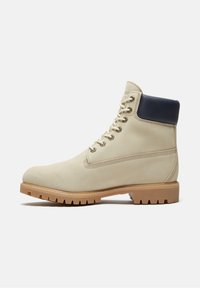 Beiger Wildleder-Arbeitsschuh mit Schnürung, schwarzem gepolstertem Kragen und dicker brauner Gummisohle mit grobem Profil, seitlich auf weißem Hintergrund gezeigt.