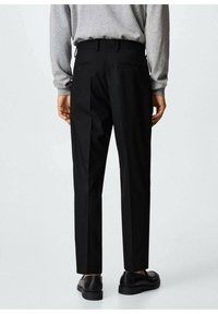 Zwarte broek met een getailleerde pasvorm, voorzien van twee achterzakken en een gladde, geweven textuur. Gecombineerd met donkere schoenen en een grijze trui.