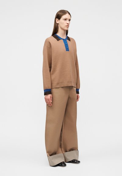 Femme debout portant un pantalon large couleur beige avec des revers à carreaux et un haut à manches longues assorti beige avec des accents de col bleus et marron.
