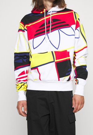 Man draagt een meerkleurige hoodie met abstracte geometrische patronen in rood, geel, zwart en blauw, en zwarte cargobroek, tegen een effen achtergrond.