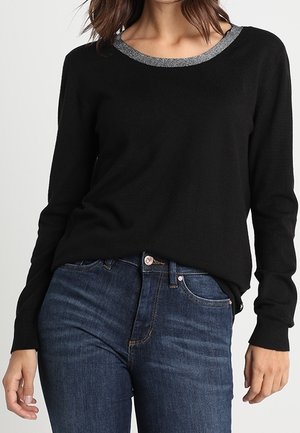 Frau trägt einen schwarzen Langarmpullover mit grauem Kragen und dunkelblaue Jeans, Hände locker an den Seiten.