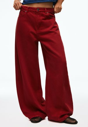 Persona che indossa pantaloni larghi e rossi abbinati a scarpe nere in mesh, mani che sistemano la vita, su sfondo bianco uniforme.