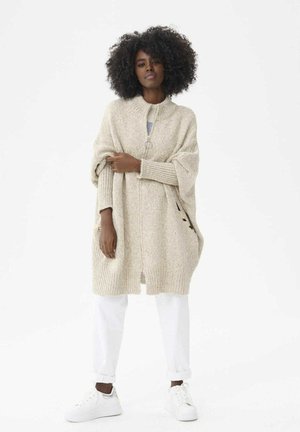 Beige gebreide cardigan met hoge kraag, open voorkant en zijzakken. Gepaard met witte broek en schoenen, wat een ontspannen silhouet creëert.