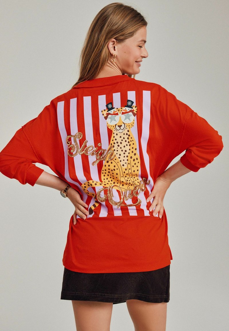 Tricou roșu supradimensionat cu dungi albe și un grafic cu ghepard. Include text strălucitor care spune "Regina Sărbătorilor" sub grafic.