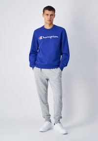 Blå sweatshirt med "Champion"-logotyp, grå sweatpants och vita sneakers. Har rund hals, ribbade mudd och elastisk midja.