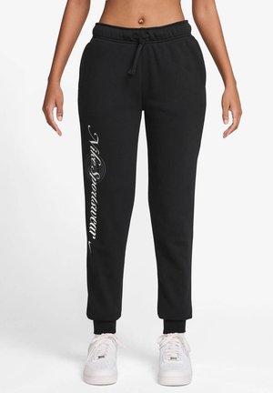 Femme portant un pantalon de survêtement Nike noir avec taille à cordon et texte de logo blanc sur la jambe gauche, associé à des baskets blanches.