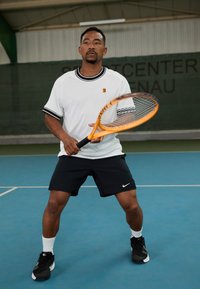 Chemise blanche à manches courtes avec des rayures noires, shorts noirs, tenant une raquette de tennis orange sur un court bleu avec un arrière-plan vert.
