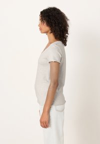 Mladá žena s kučeravými tmavými vlasmi, oblečená v svetlosivej tričku s krátkym rukávom a svetlomodrých džínsach, stojaca v profile na bielom pozadí.