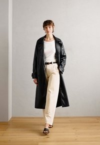 Trench coat in pelle nera sopra una maglietta bianca, abbinato a pantaloni beige e sandali neri a scivolo; tessuto liscio, design classico, cappotto lungo fino al ginocchio.