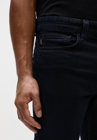 Jeans de mezclilla azul oscuro con un diseño ajustado, que presentan un bolsillo frontal y una etiqueta de logo visible en naranja. Textura suave, cinturilla estándar.