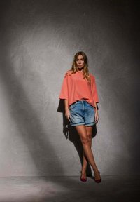 Un haut corail oversize, un short en denim et des talons roses. Le modèle se tient contre un fond gris texturé, projetant une ombre subtile.