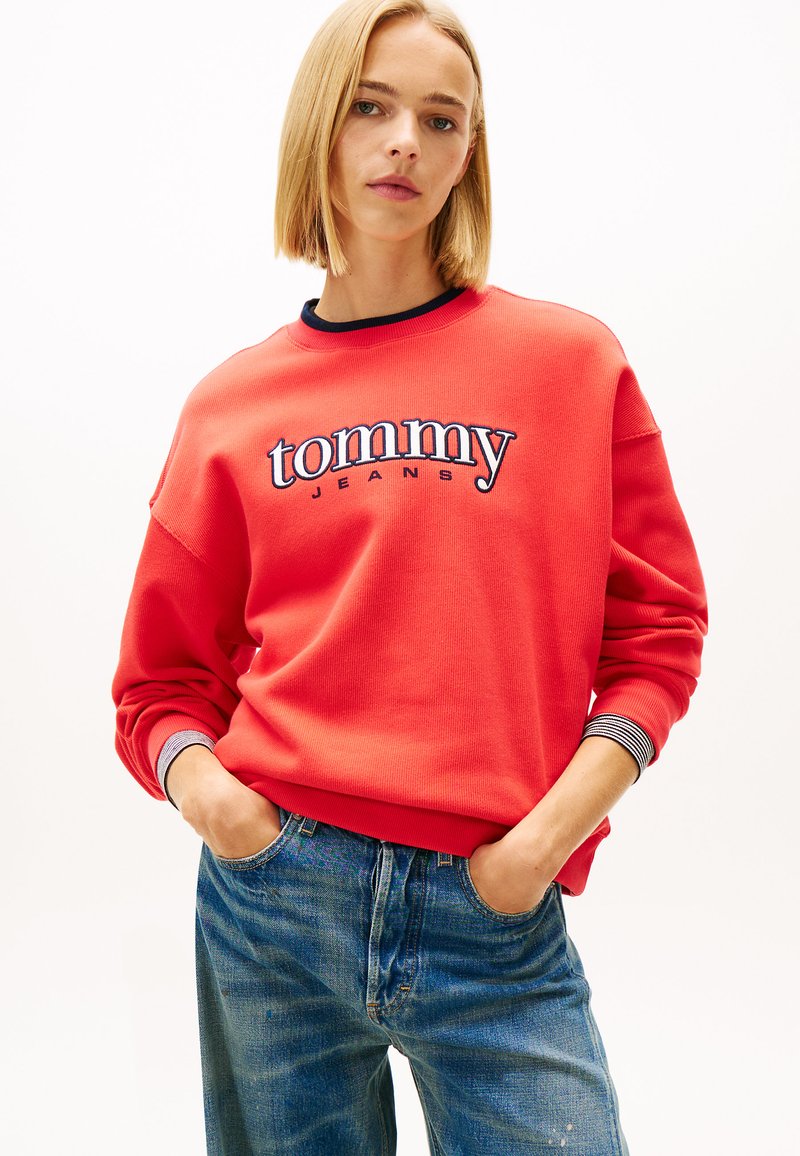 Červená mikina s rebrovanou textúrou, s modrým logom "tommy" a nápisom "JEANS" v námorníckej modrej. Manžety majú pruhovaný vzor v námorníckej a bielej farbe. V páre s modrými džínsami.