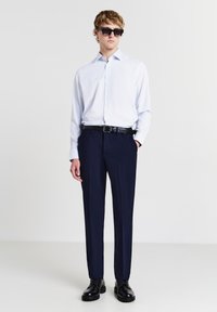 Giovane uomo con occhiali da sole neri, camicia a maniche lunghe azzurro chiaro, pantaloni navy scuri, cintura nera e scarpe eleganti nere, in piedi con una mano in tasca.