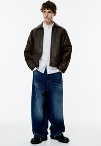 Veste en simili cuir marron avec un col, portée sur une chemise blanche à boutons. Un jean en denim bleu à jambes larges et des chaussures noires épaisses complètent le look.