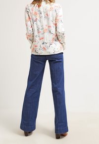 Bloes met bloemenpatroon in crême met roze en blauwe bloemen, gecombineerd met donkerblauwe wijde jeans en bruine hakken.