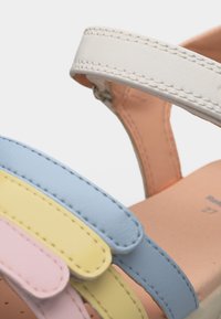 Κοντινό πλάνο ενός σανδαλιού με λευκό λουράκι αστραγάλου και παστέλ ροζ, κίτρινα και μπλε λουράκια στα δάχτυλα, που δείχνει λεπτομερή ραφή σε καφέ πάτο.