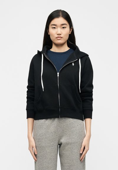 Νεαρή γυναίκα που φορά μαύρο φούτερ με φερμουάρ και λευκά κορδόνια, μπλε-ναυτικό πουκάμισο και γκρι φόρμα, στέκεται μπροστά σε λευκό φόντο.