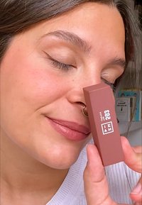 Matte brunt läppstiftstub som hålls nära ansiktet, med en elegant rektangulär form, vit textetikett och en rundad spets på applikatorn.