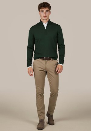 Man kijkt recht vooruit, draagt een donkergroene trui met kwartrits, wit overhemd, beige broek, bruine riem en bruine schoenen tegen een egale achtergrond.