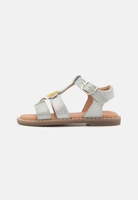 Silberne glitzernde Sandalen mit drei breiten Riemen, einem seitlichen Schnallenverschluss und einem dekorativen gelben Blumenakzent auf der Vorderseite. Braune Sohle.