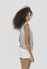 Szürke ujjatlan felső fehér geometriai mintákkal, fehér rövidnadrággal párosítva. Az anyag puha és laza szabású. A modellnek dús haja van.