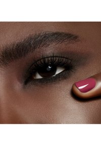 Close-up of dark brown eye with long eyelashes and smoky eyeshadow, with a finger showing glossy dark pink nail polish near the cheek.
