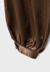 Manica di camicetta in raso marrone con polsino abbottonato, tessuto arricciato che crea un effetto gonfio su uno sfondo chiaro.