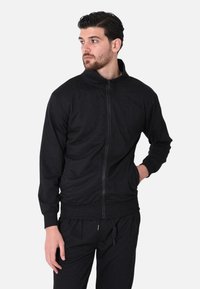 Giacca nera con zip, realizzata in tessuto morbido, con colletto alto e polsini a coste, indossata con pantaloni neri dotati di tasche laterali.