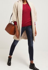 Crème getextureerde open cardigan over een rood geribd langslijfshirt, gecombineerd met donkere skinny jeans en zwarte enkellaarsjes, en een bruine handtas.