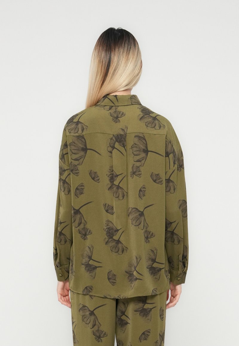 Person mit langen blonden Haaren, die eine olivgrüne Bluse und Hose mit dunklem Blumendruck trägt, steht vor einer weißen Wand.