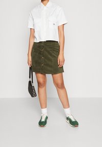 Chemise blanche à manches courtes avec deux poches poitrine, mini-jupe en velours côtelé vert avec boutons sur le devant, associée à des baskets vertes et un sac noir à motifs.